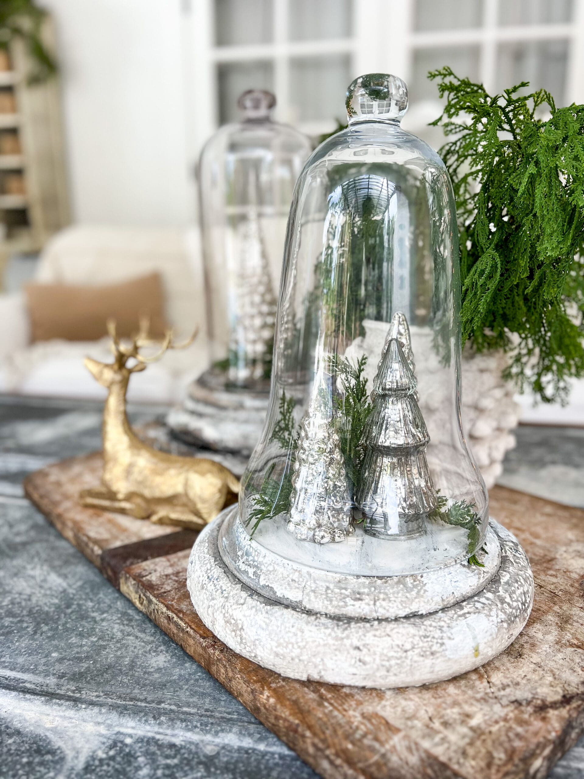 a cloche with mercury glass trees inside on top of a wooden base next to a unique brass deer with a small tree behind it