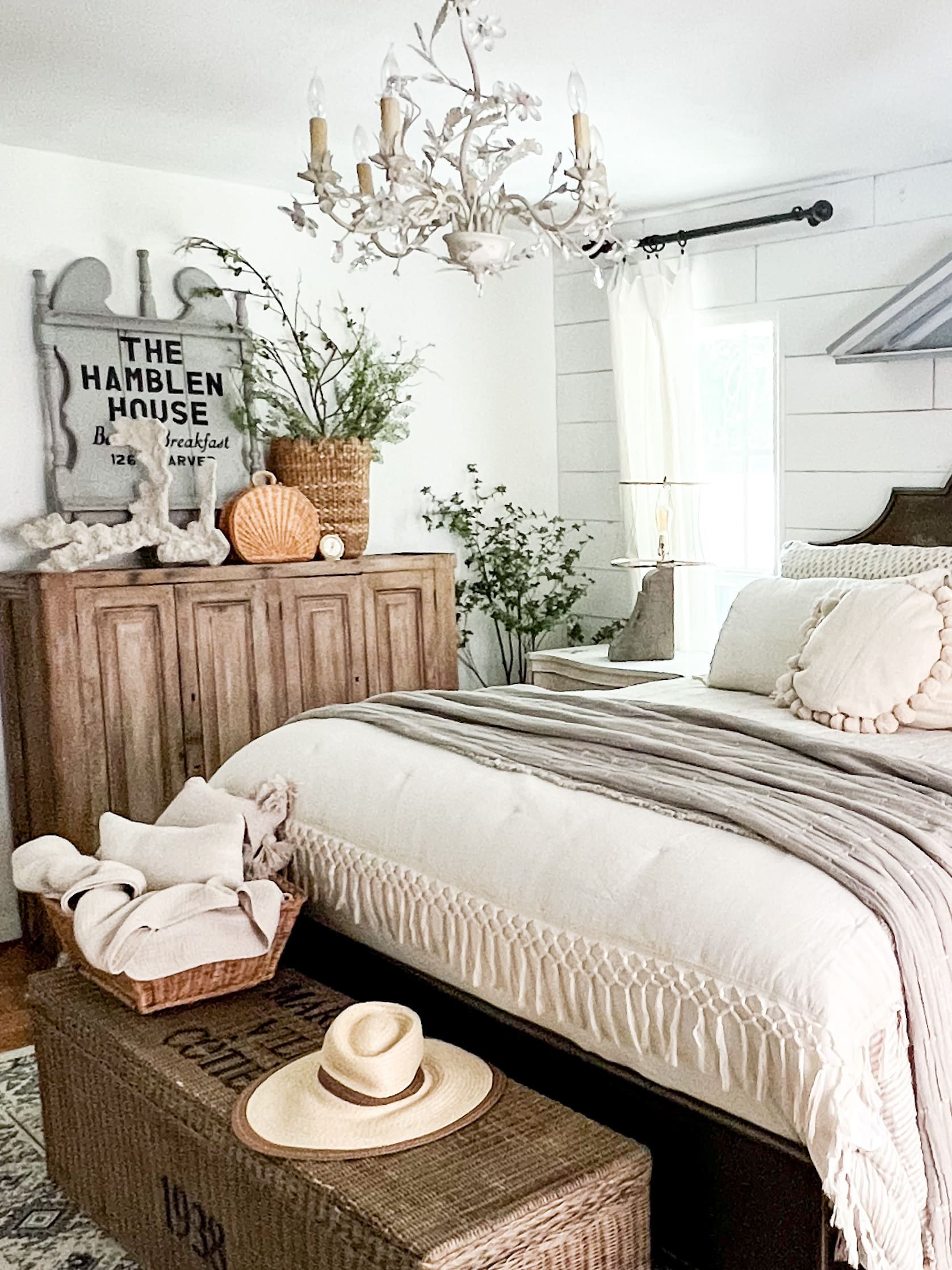 summer bedroom styled with a wooden dresser with old gray sign and coral pieces, a big woven basket at the foot of metal bed frame styled with vintage hat