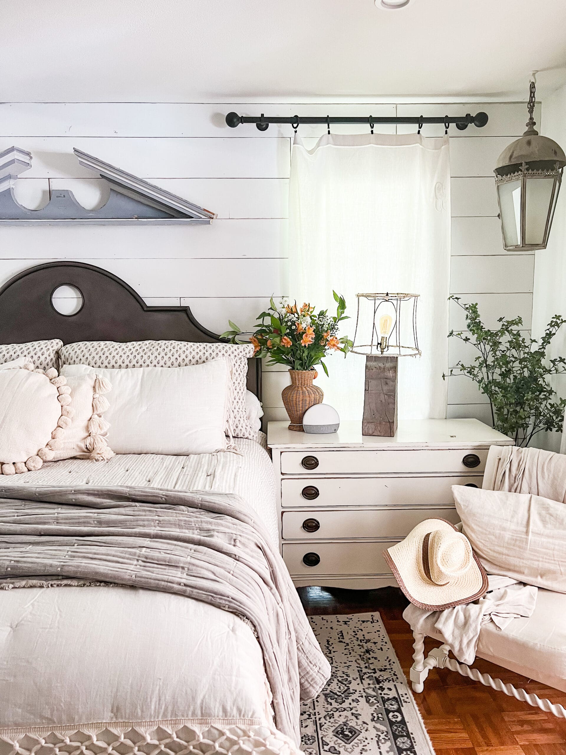 cute white dresser styled with fresh flowers in woven vase and a lamp made out of salvaged wood, next to a side chair with cute throw blanket and pillow