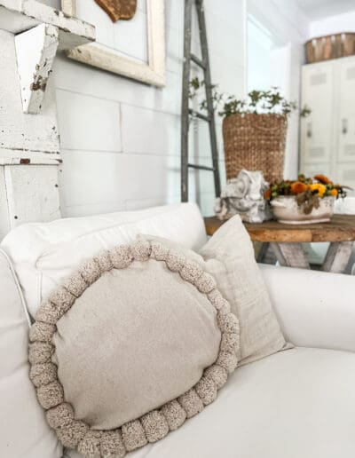 Round grayish-white pillow with pom-poms on white sectional and fall bouquet in background