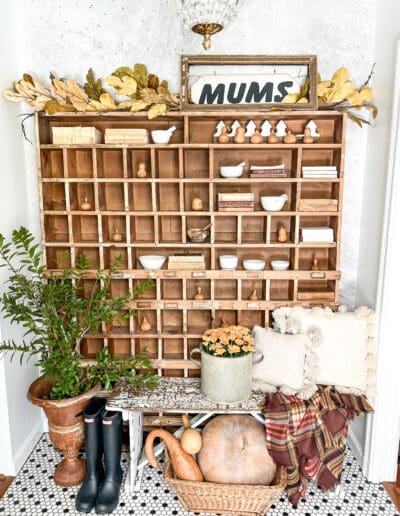 Wooden cubbies in entryway with small gourds and mums sign, with white bench and plaid throw in front.