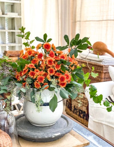 Orange fall blooms with fresh tree trimmings in arrangement in vintage white pot with handle.
