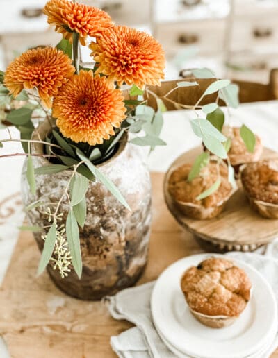 Orange Mums with eucalyptus filler in vintage crock, with fresh muffins in the background.
