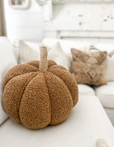 Fuzzy, brown, pumpkin-shaped throw pillow on a white couch with other neutral pillows in the background