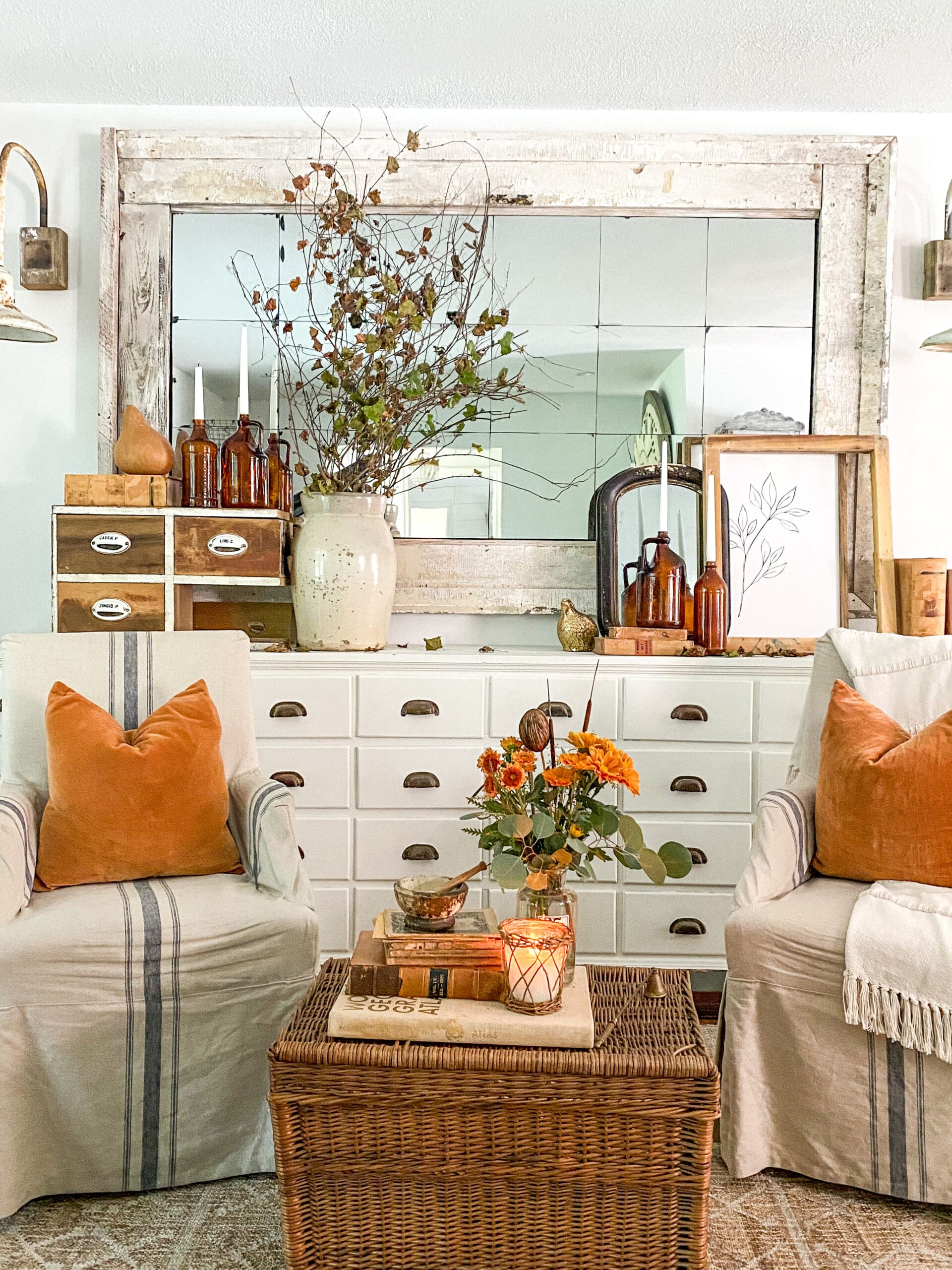 White vintage apothecary, large mirror, and sitting chairs styled for fall with rust tones, vintage frames, and fresh flowers
