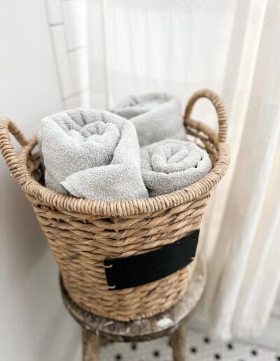 Sturdy wicker basket holds rolled-up white towels on vintage wooden stool, complete with decades-old paint spots.