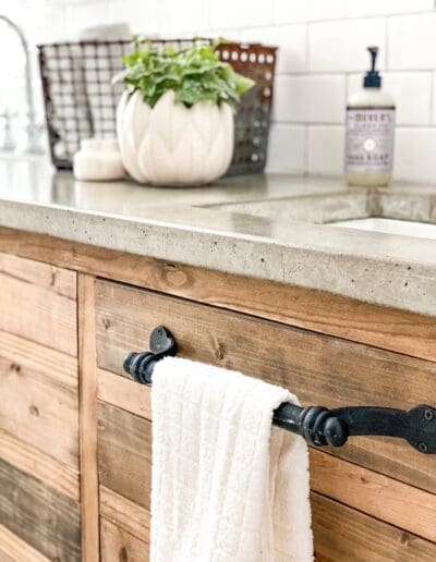 Black towel bar screwed into one of the vanity cabinet doors. Simple white towel hangs down in front of image to match the rest of the wood/black/white/concrete them.
