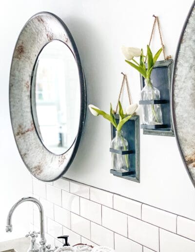 Neatly installed white subway tile form a backsplash behind the two matching sinks. Two pieces of wall art with faux tulips hang on the wall above between two matching galvanized oval mirrors.
