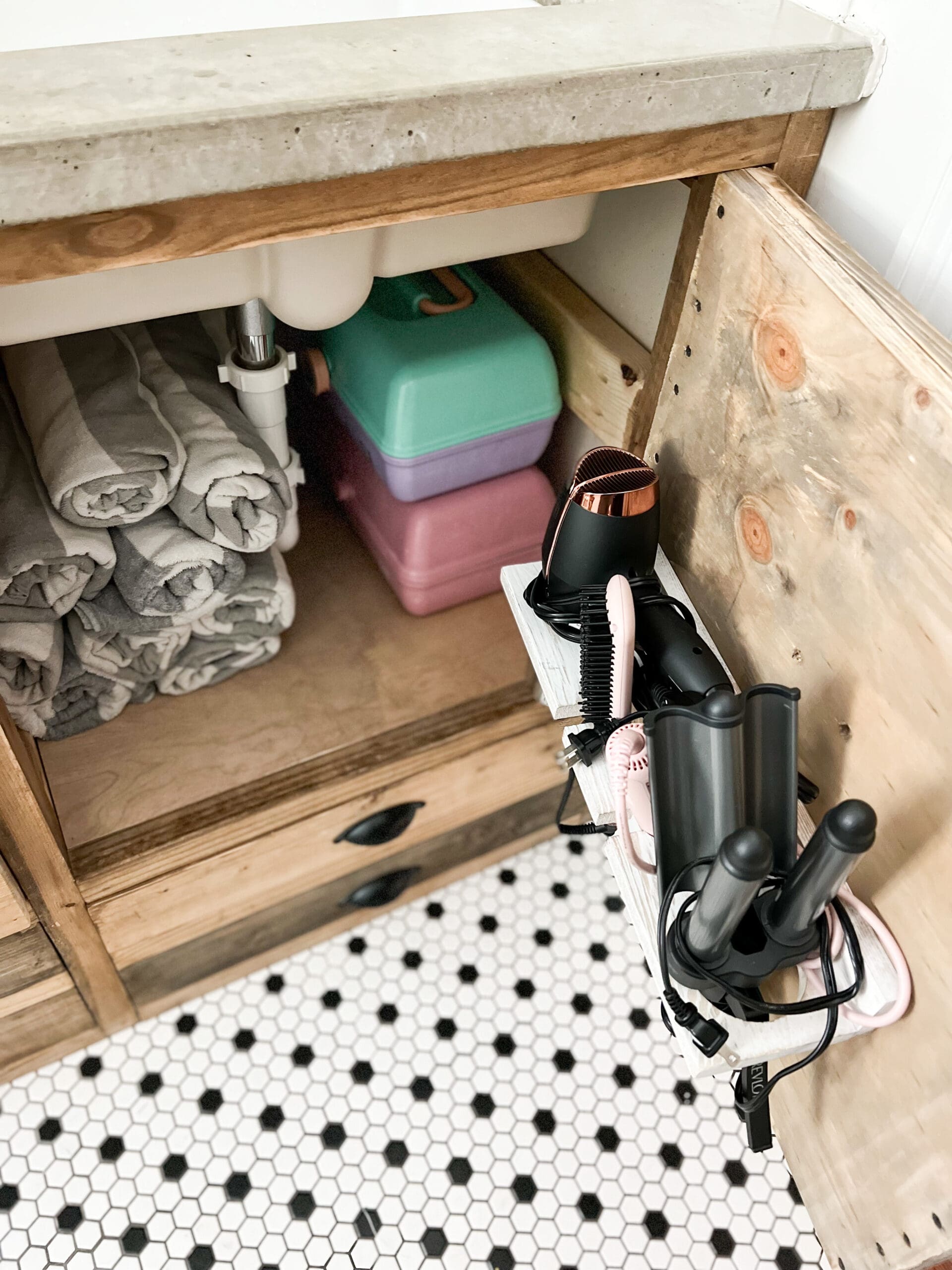 With cabinet door open, neatly rolled up gray and white pools towels are seen. Hair dryer and other accessories fit in a white holder screwed to the back of the cabinet door.
