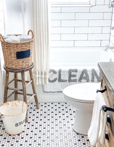 Showing the white shower curtain with white bathmat & gray "CLEAN" letters hung neatly over bathtub. A wicker basket of towels sits on a vintage wooden stool besides the tub.