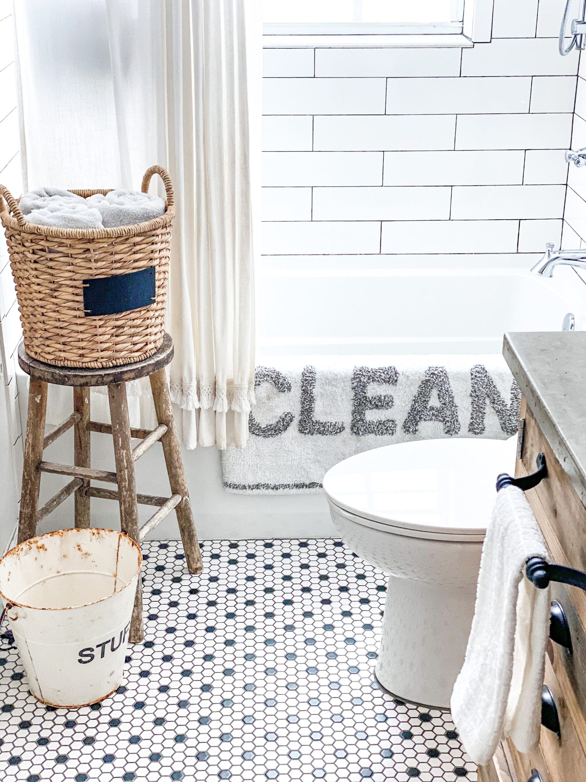 Showing the white shower curtain with white bathmat & gray "CLEAN" letters hung neatly over bathtub. A wicker basket of towels sits on a vintage wooden stool besides the tub.