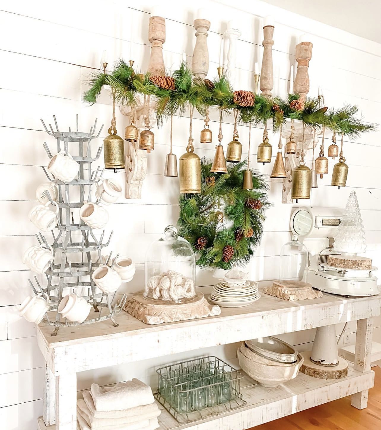 side view of a chippy white table styled for Christmas with glass cloches, Christmas greenery, golden bells hung above, and a large white scale with a glass Christmas tree on top
