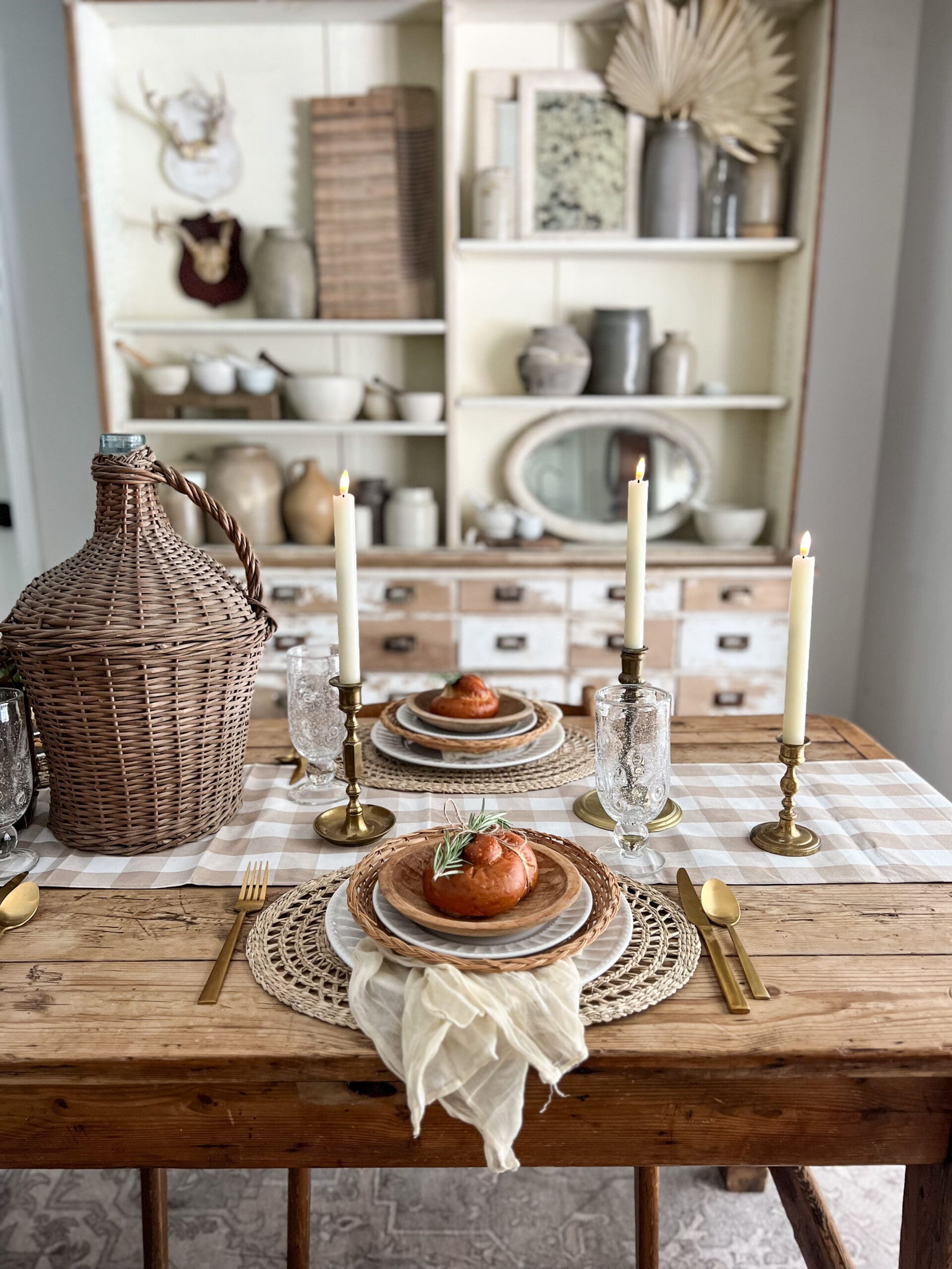 Close up of a fall place setting with gold flatware and candlesticks, woven chargers and freshly baked rolls