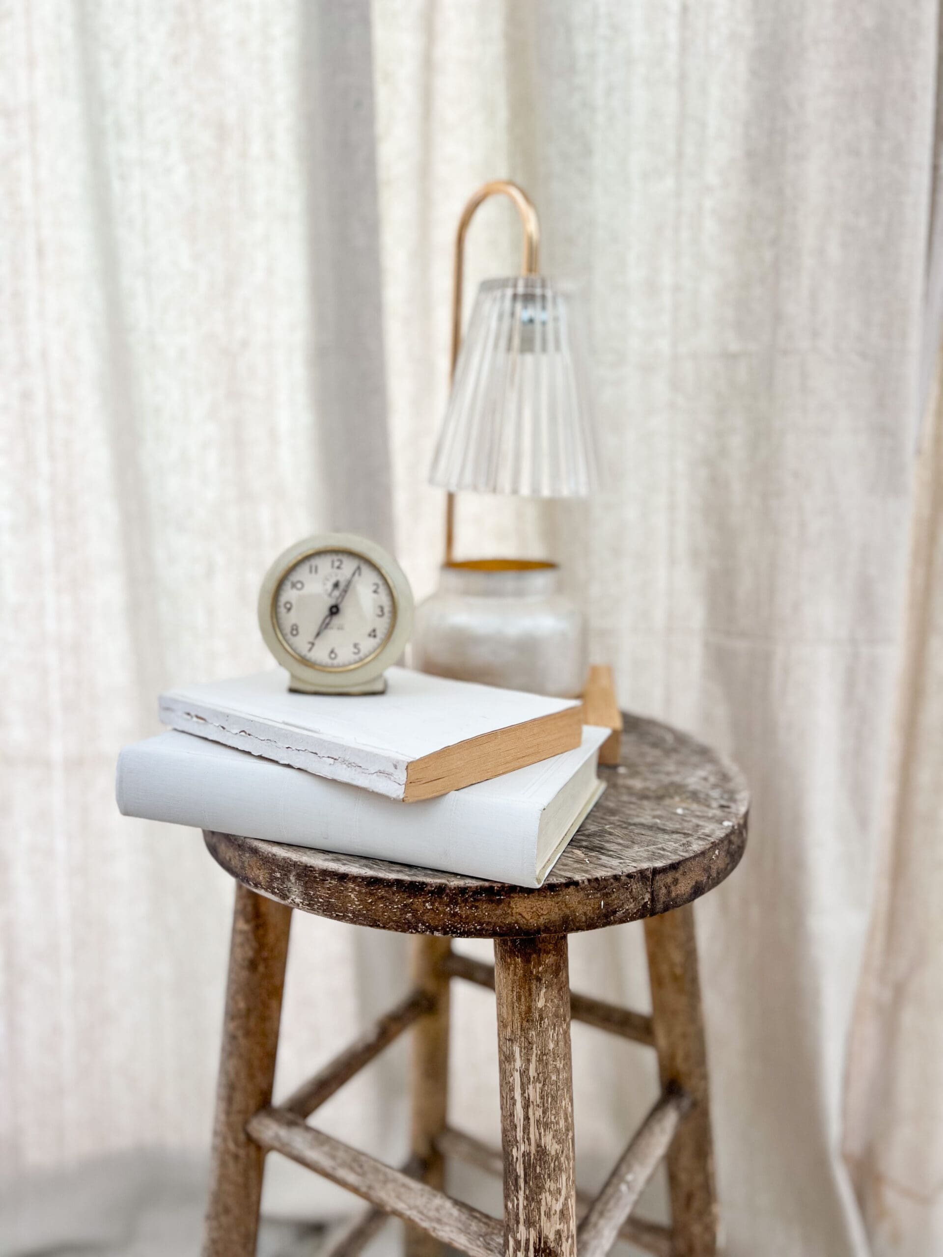White vintage books, a small white clock, and a candle warmer sitting on a round wooden stool