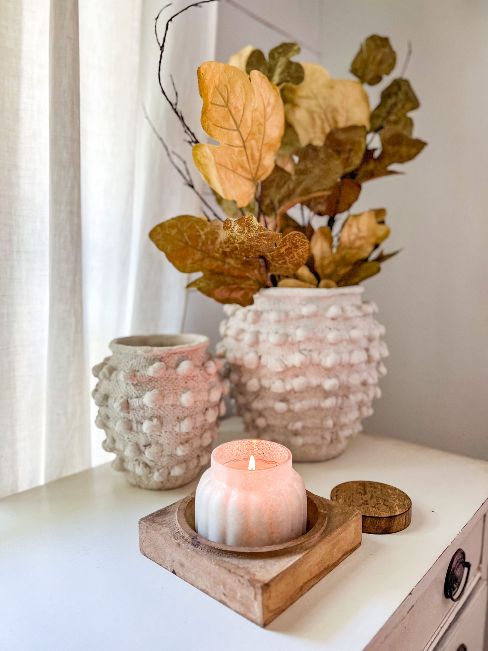 vignette styled with two white textured vases holding dried leaves, and a cozy candle on a wooden base