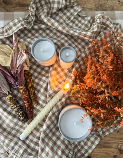 A brown gingham cloth with gray candles, corn husks, fall faux greenery, and a battery-operated candlestick