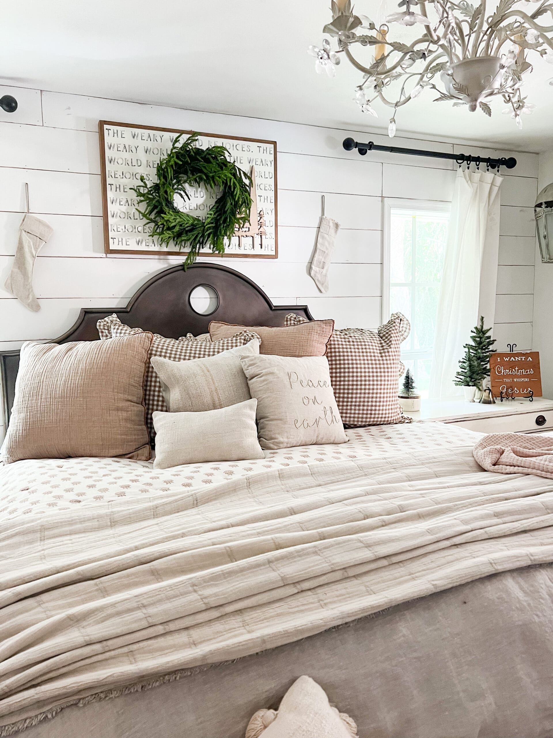 view of a bed with neutral bedding and a christmas pillow with a Christmas quote on a sign above the bed, and a wreath hanging over the sign