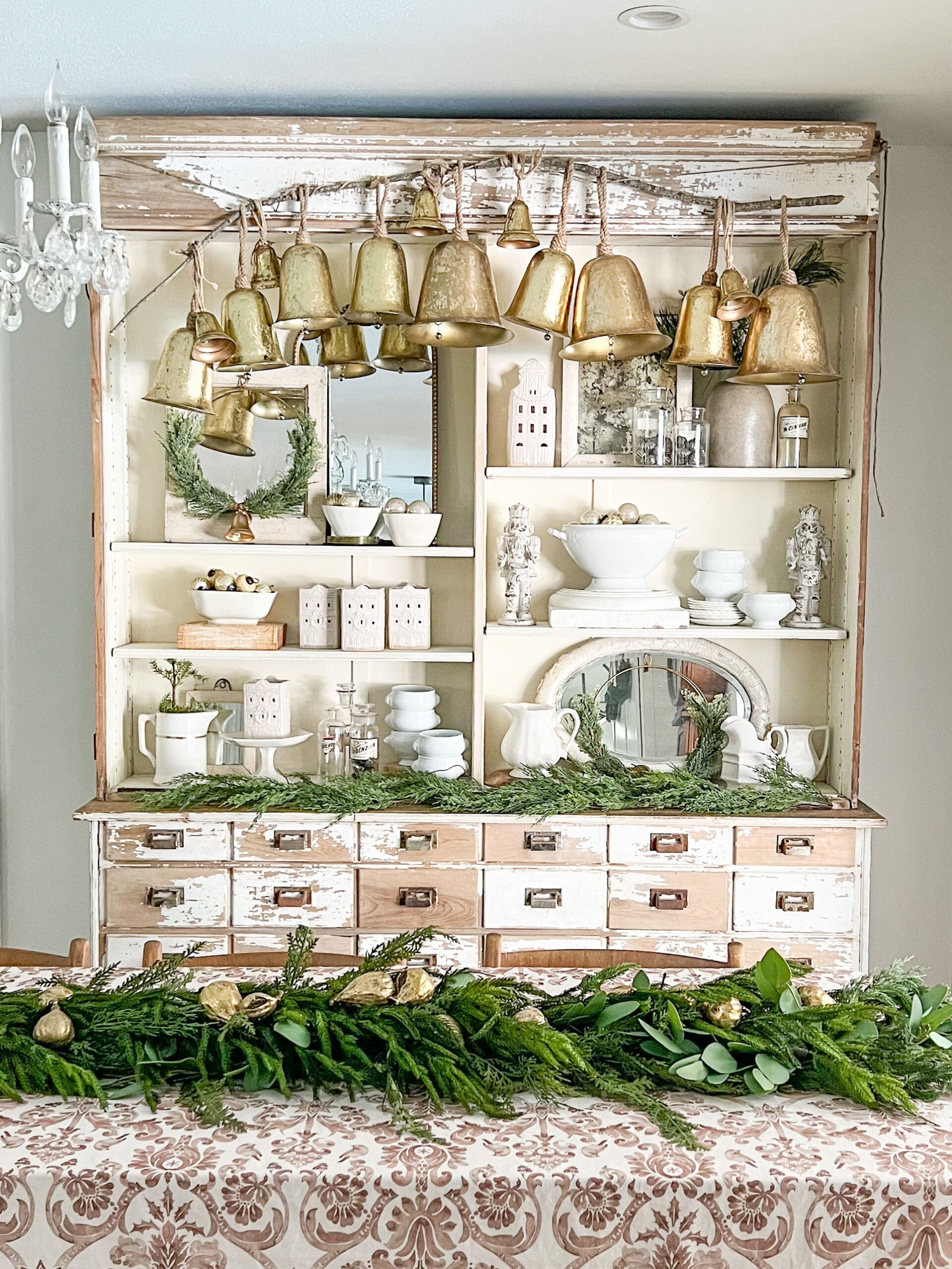 chippy white apothecary styled with large golden bells hung from the top and different elements of Christmas decor styled on the shelves