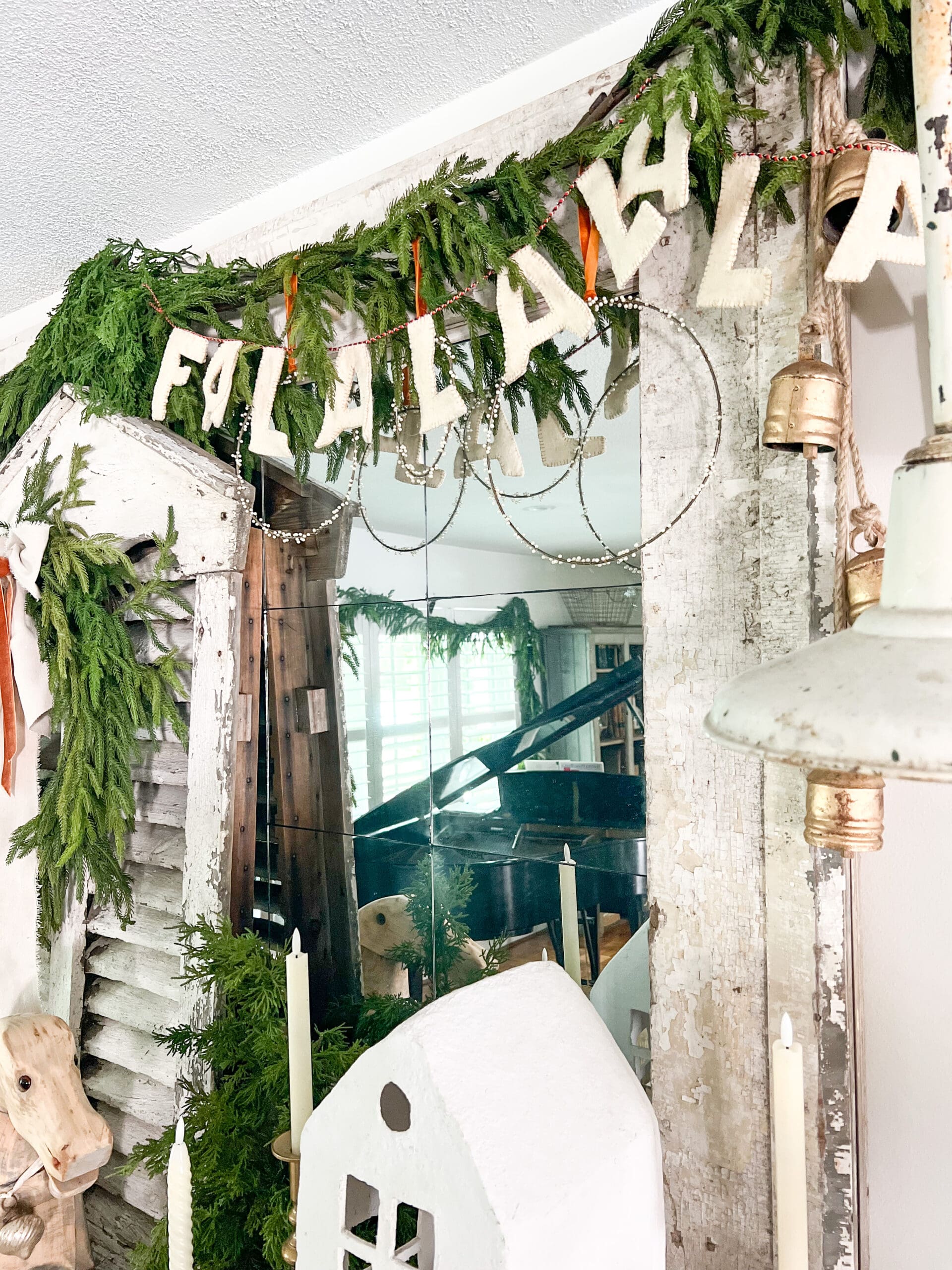 "Fa la la" garland hung on a large mirror with Christmas greenery framing the mirror and golden bells hanging from the edges