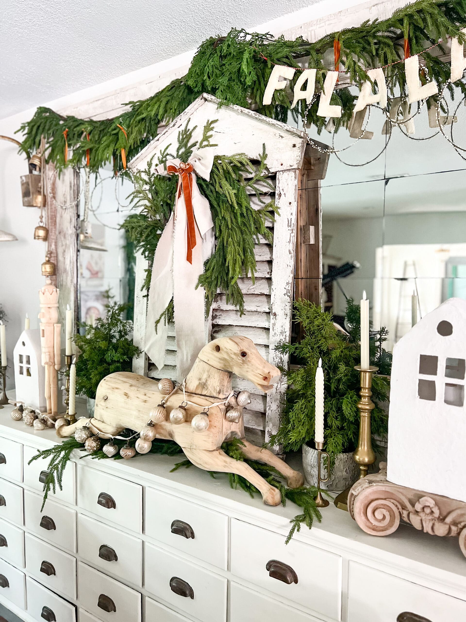 white apothecary styled with a wooden rocking horse, a large mirror with a "fa la la" sign, and Christmas greenery hung from it