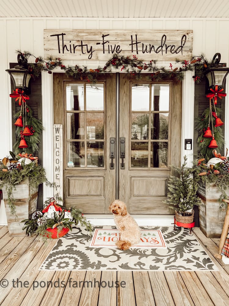 two wooden double front doors styled with Christmas greenery and black lanterns on either side of the door and a dog named Rudy posed in front of the door