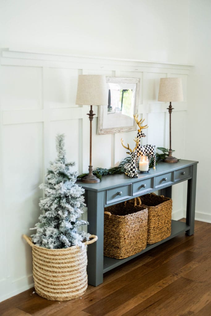 black console table with lamps and Christmas decor on top and a medium height Christmas tree in a basket beside the table