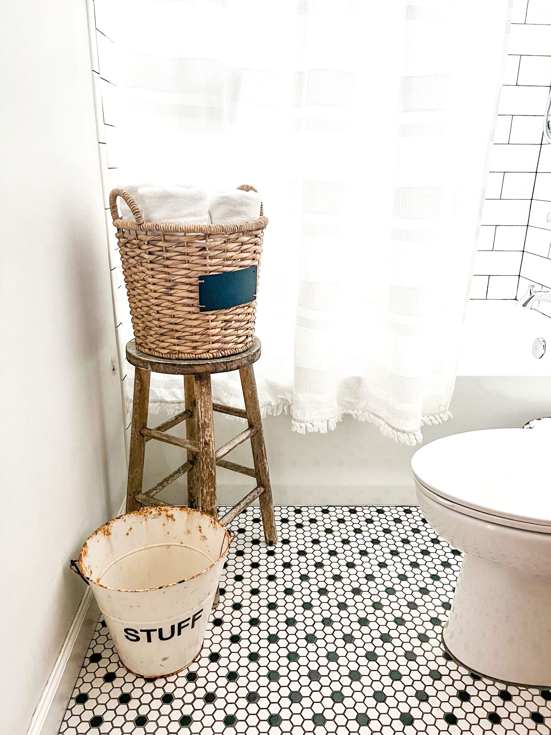 a wood stool with a basket on top holding towels next to a shower and a small metal bucket next to it for decor