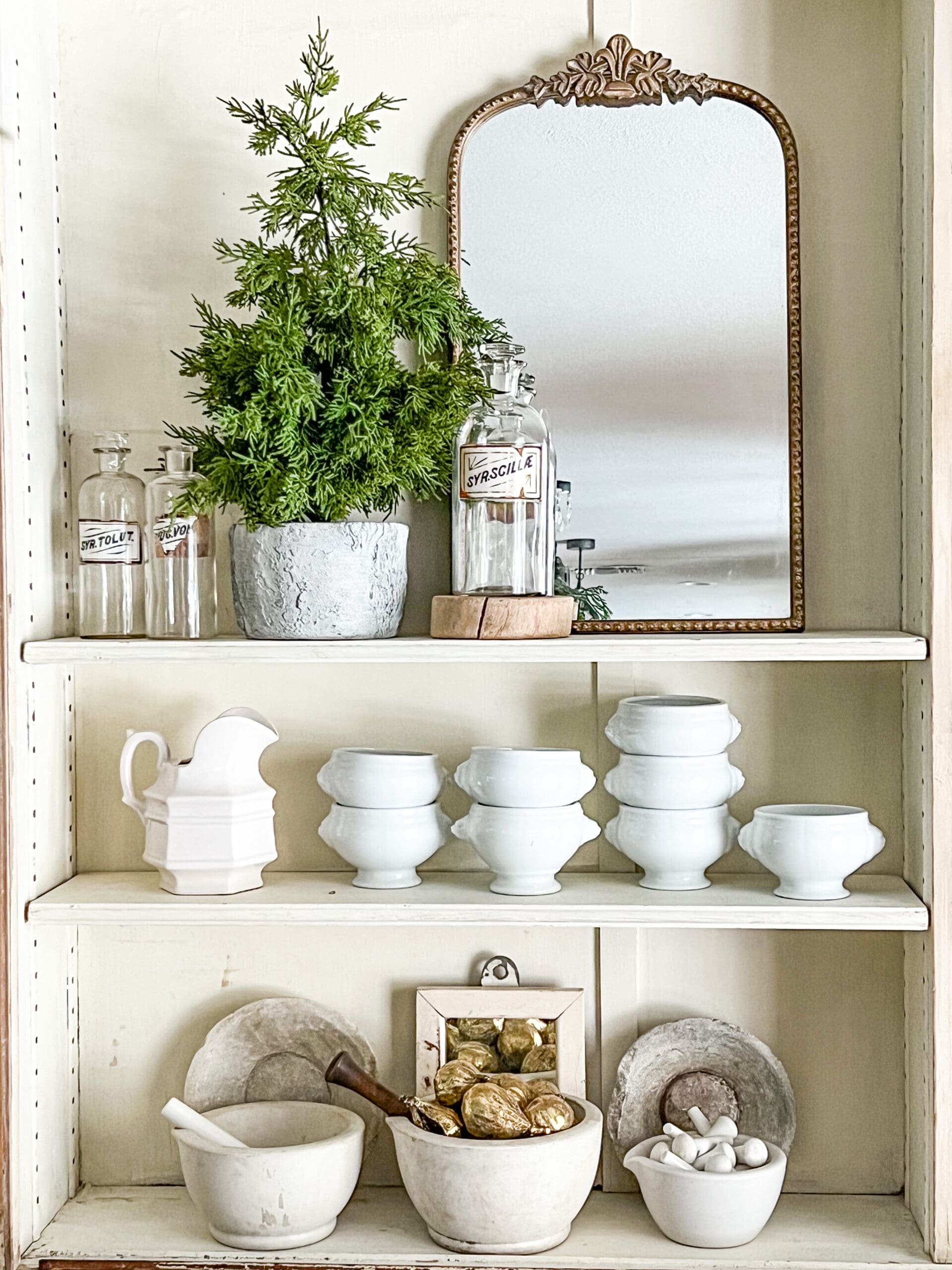 shelving styled with a medium-sized decorative tree, a gold mirror, several mortar and pestles with decorative accents inside, and ironstone