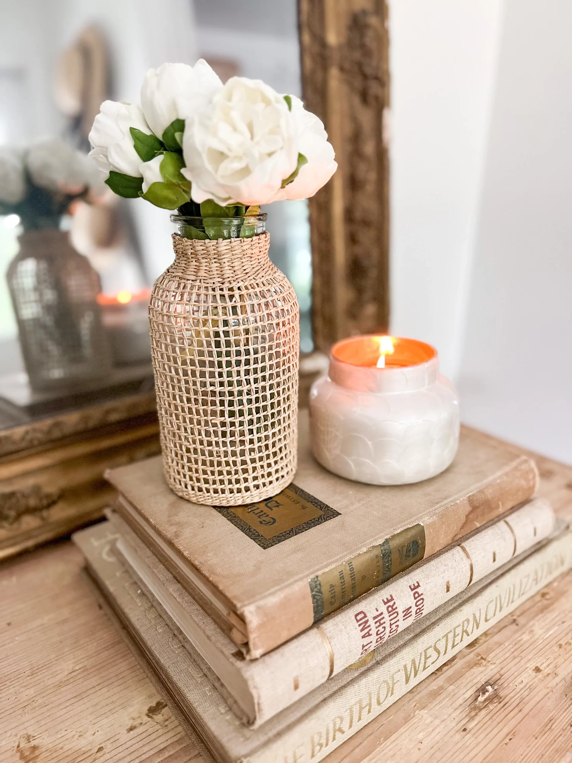 small woven vase with faux peonies next to a small candle on top of a stack of vintage books